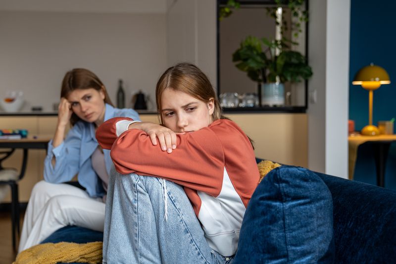The photo shows an adolescent girl sitting on a sofa with her mother in the background. The girl is sitting on profile while she looks towards the camera. She has her knees pulled up to her body so she looks upset while her mother sits at the other end of the sofa and looks at her with a worried or frustrated look. The scene is set in a living room.