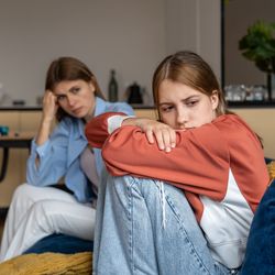 The photo shows an adolescent girl sitting on a sofa with her mother in the background. The girl is sitting on profile while she looks towards the camera. She has her knees pulled up to her body so she looks upset while her mother sits at the other end of the sofa and looks at her with a worried or frustrated look. The scene is set in a living room.