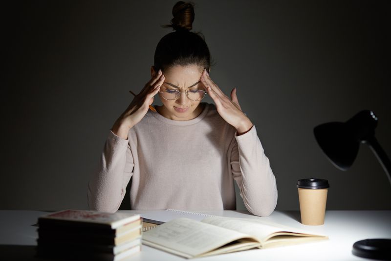 woman wearing glasses sitting at desk looking stressed, with books, coffee cup and lamp