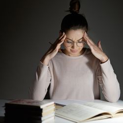 woman wearing glasses sitting at desk looking stressed, with books, coffee cup and lamp