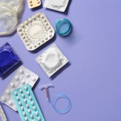 purple background with various contraceptives arranged towards the left hand side: pills, condoms, and an intrauterine device.