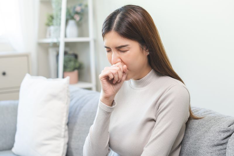 woman with long hair wearing beige sweater sitting on a grey sofa and coughing
