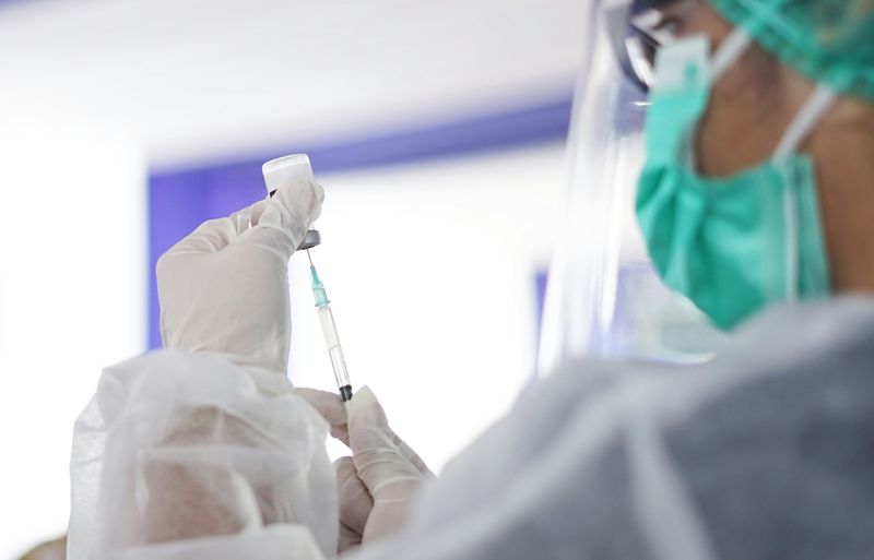 medical professional in green surgical mask, gown, face shield and gloves draws up vaccine into syringe