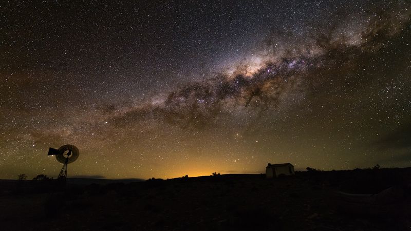 A night view. A windmill and a house are seen as silhouttes. IN the sky the milky way dominates the whole sky.