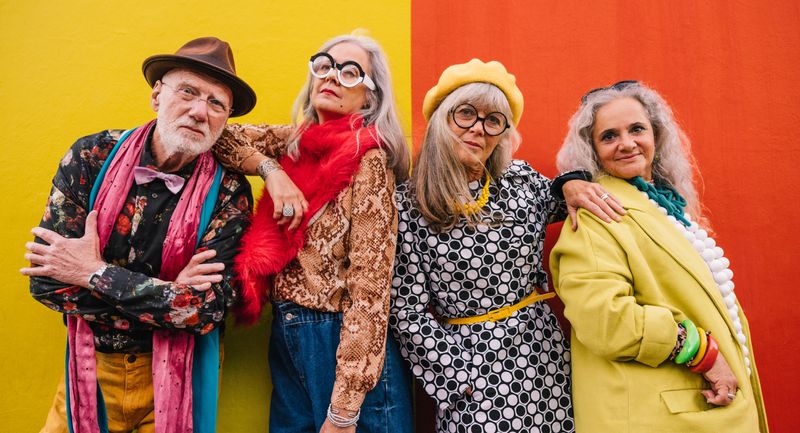 Four older people, a man and three women, stand in a line and are posing and leaning on one another. They are all dressed in colourful eccentric clothing and the background is divided between yellow (on the left) and red (on the right).