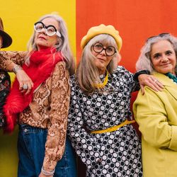 Four older people, a man and three women, stand in a line and are posing and leaning on one another. They are all dressed in colourful eccentric clothing and the background is divided between yellow (on the left) and red (on the right).