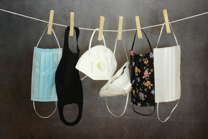  different types of face coverings, including cloth and surgical masks and disposable respirators, hanging on a washing line with wooden pegs against a grey background