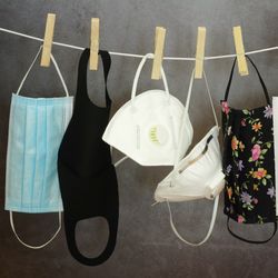  different types of face coverings, including cloth and surgical masks and disposable respirators, hanging on a washing line with wooden pegs against a grey background