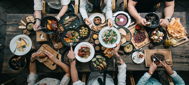 aerial view of a dinner party with lots of different dishes being passed between six people, seated three on either side of a long wooden table