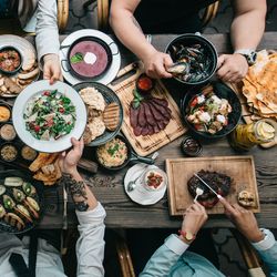 aerial view of a dinner party with lots of different dishes being passed between six people, seated three on either side of a long wooden table