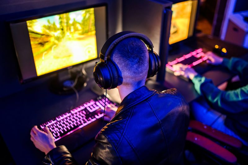 A man wearing a leather coat and headphones is sitting with his back to the camera. He is in dark space playing a videogame while typing at a keyboard that has a pink illumination running through the keys. 