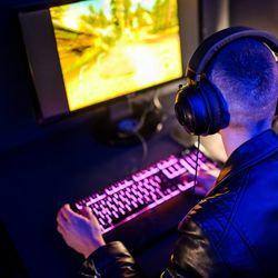 A man wearing a leather coat and headphones is sitting with his back to the camera. He is in dark space playing a videogame while typing at a keyboard that has a pink illumination running through the keys. 
