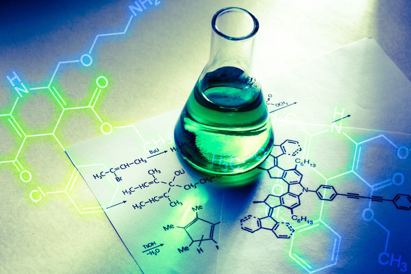 Conical flask filled with green liquid on top of a sheet of paper with chemical reactions written on
