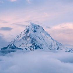 A photo of a mountain top peaking out above the clouds. The mountain top is wedge-shapes and covered in snow with a few patches of rock showing through. The sky behind is pale, illuminated by the morning light. 