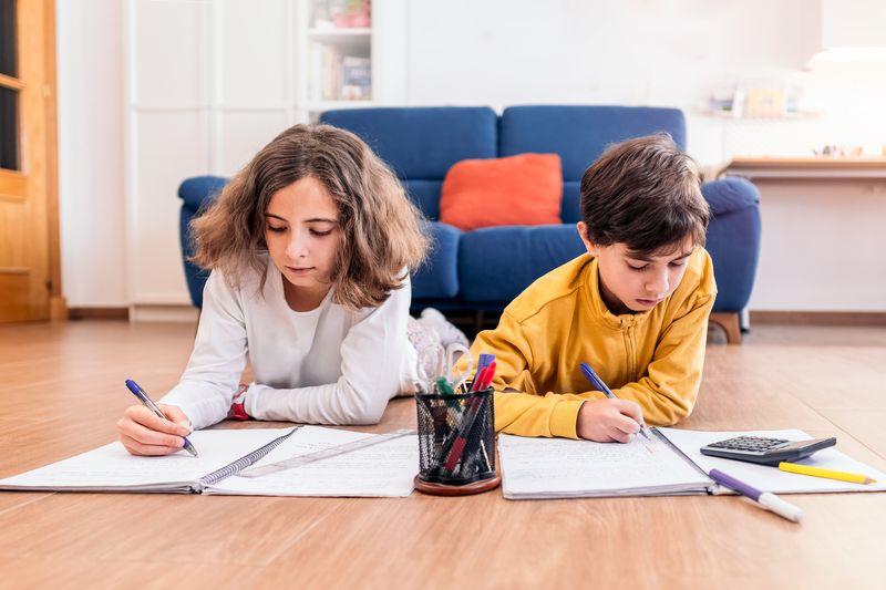 two kids lying on the floor on their stomachs doing homework; one has a calculator and there's a pot of pens between them; there's a blue sofa in the background and the floor is light wood