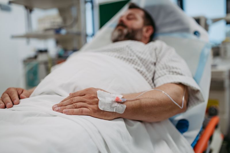 man with beard lying in hospital bed with a cannula in the back of his hand