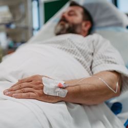 man with beard lying in hospital bed with a cannula in the back of his hand