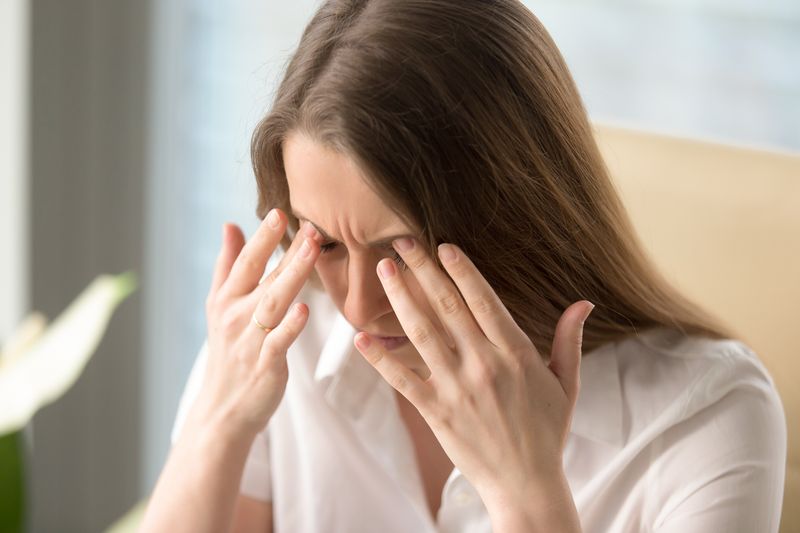 Woman wearing white shirt pressing her fingers around her eyes as though in pain.