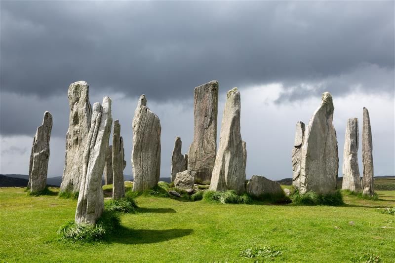 A photo of the standing stones on the the Isle of Lewis and Harris, in the Outer Hebrides. The stones are arranged in a lush green field and the photo shoes a dark and story sky. Some of the stones have weathered into points whereas others still retain a more regular shape. 