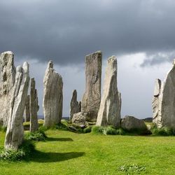 A photo of the standing stones on the the Isle of Lewis and Harris, in the Outer Hebrides. The stones are arranged in a lush green field and the photo shoes a dark and story sky. Some of the stones have weathered into points whereas others still retain a more regular shape. 