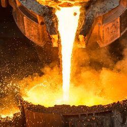 A ladle pouring molten metal that looks orange like it's on fire into another poth, sparks are flying up all around. 