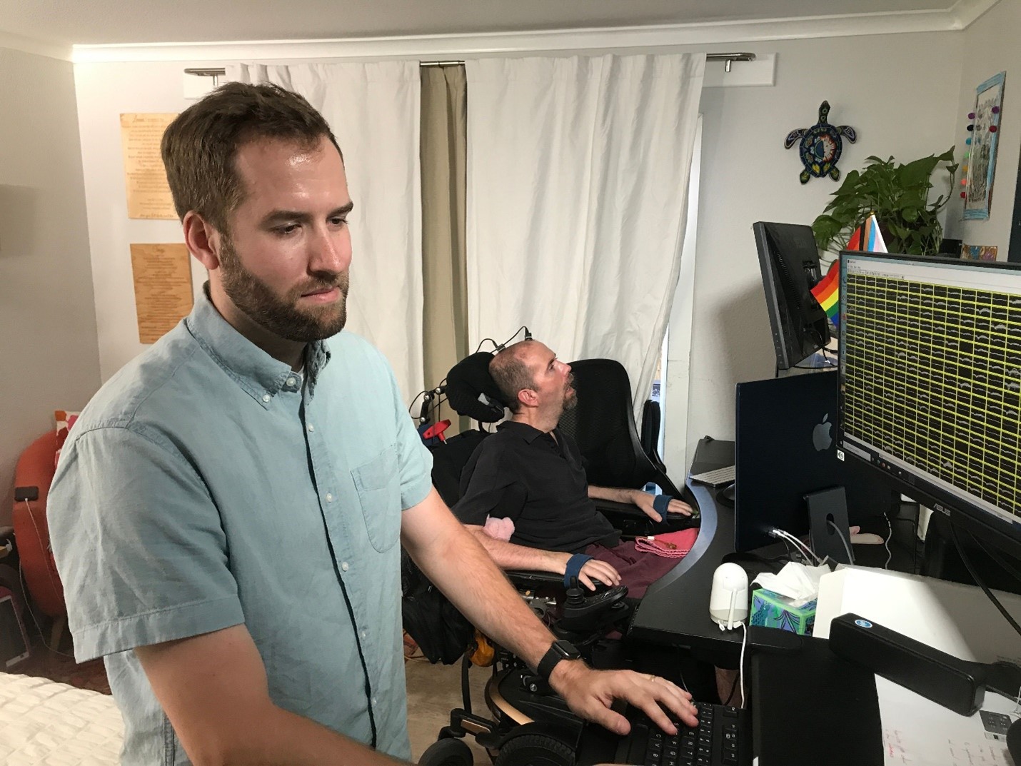 Postdoctoral scholar and lead author of the study Nicholas Card getting the BCI system ready. Card is in the foreground, a white man with brown hair and bear wearing a pale blue short sleeved shirt. He is standing in front of a computer. In the background Casey Harrell, a white man who uses an electric wheelchair, is positioned in front of another computer monitor.