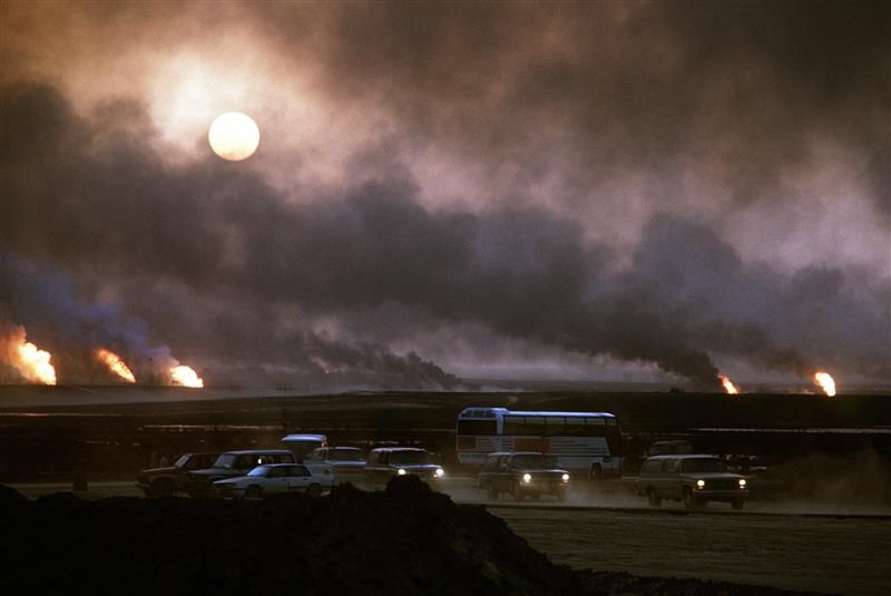 The photo shows a sky filled with smoke as the oil wells burnt in Kuwait in 1991. The sun can be seen trying to penetrate the cloud of smoke. The horizon of the image shows five fires in the distance, three on the left hand side and two on the right, which are the source of the towers of smoke. In the foreground cars and vehicles can be seen with their headlights on despite it being daytime. 