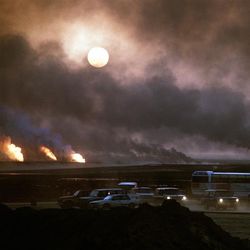 The photo shows a sky filled with smoke as the oil wells burnt in Kuwait in 1991. The sun can be seen trying to penetrate the cloud of smoke. The horizon of the image shows five fires in the distance, three on the left hand side and two on the right, which are the source of the towers of smoke. In the foreground cars and vehicles can be seen with their headlights on despite it being daytime. 