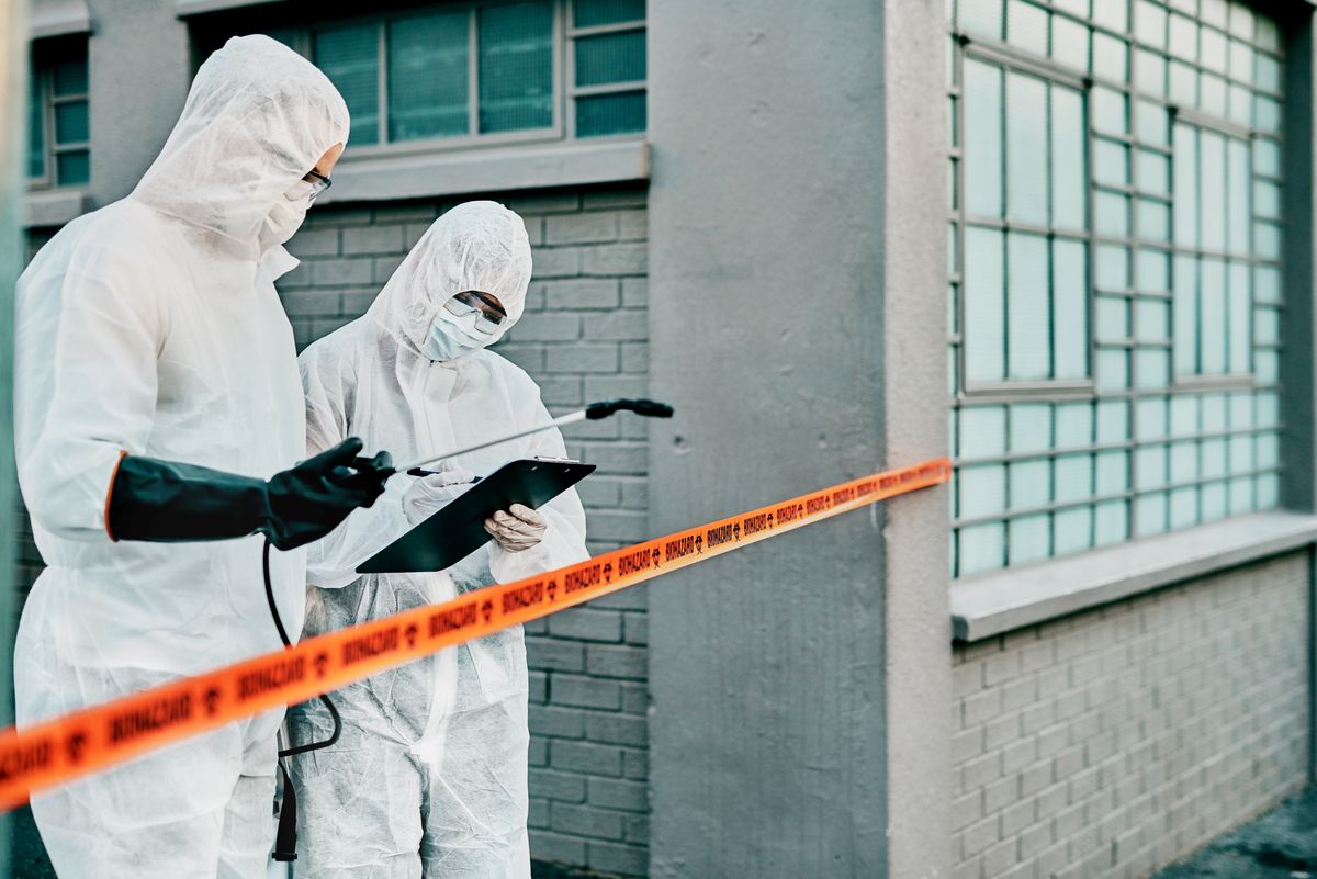 two people in HazMat suits looking at a clipboard, standing behind orange "biohazard" tape