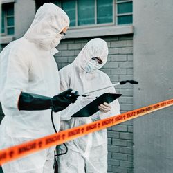 two people in HazMat suits looking at a clipboard, standing behind orange "biohazard" tape