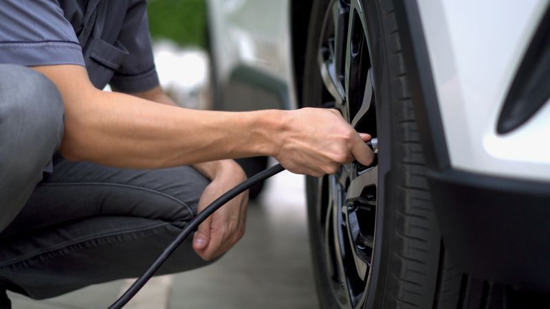 Person in jeans and blue T-shirt crouching down, holding a black rubber hose attached to a car tire