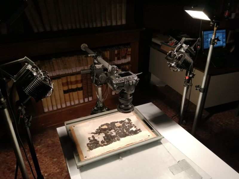 blackened scroll fragments being photographed under visible lights; the fragments are laid on a light background with a camera suspended above and lights on them; in the background are bookshelves and a desk with a computer