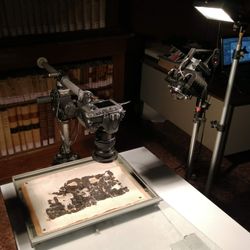 blackened scroll fragments being photographed under visible lights; the fragments are laid on a light background with a camera suspended above and lights on them; in the background are bookshelves and a desk with a computer