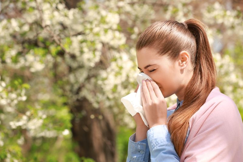 girl blowing nose into a tissue, trees blooming with flowers in the background