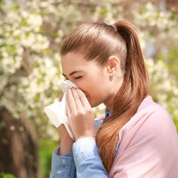 girl blowing nose into a tissue, trees blooming with flowers in the background
