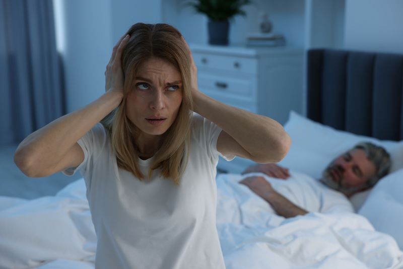 woman covering ears while man snores in bed in background