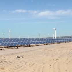 solar panels laid out in a diagonal line across the desert with wind turbines visible in the background