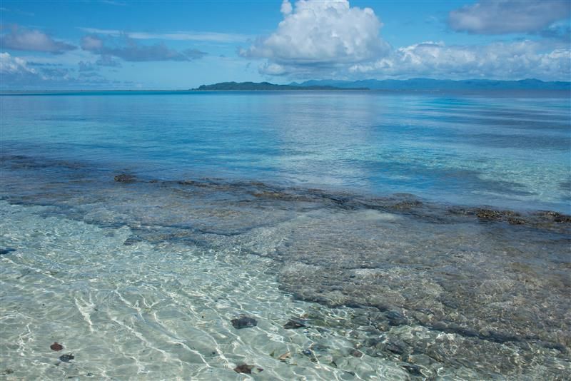 A photo taken from the shore of Melanesia, Makira-Ulawa Province, Solomon Islands. The image shows clear water and coral in the foreground and then a small island in the distance. The sky is mostly clear with a few large white clouds.  