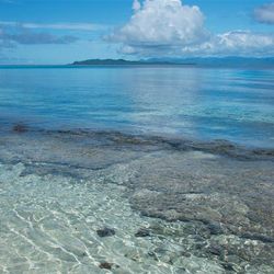 A photo taken from the shore of Melanesia, Makira-Ulawa Province, Solomon Islands. The image shows clear water and coral in the foreground and then a small island in the distance. The sky is mostly clear with a few large white clouds.  