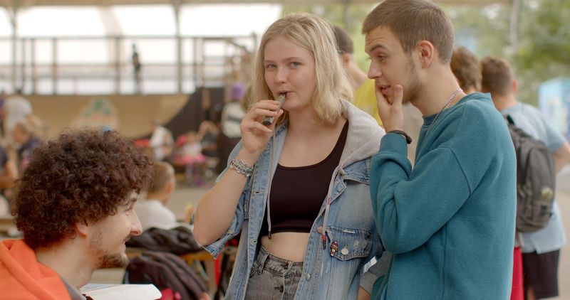 Photo of three young people, girl in the middle is holding a vape up to her mouth while the two boys either side of her are chatting