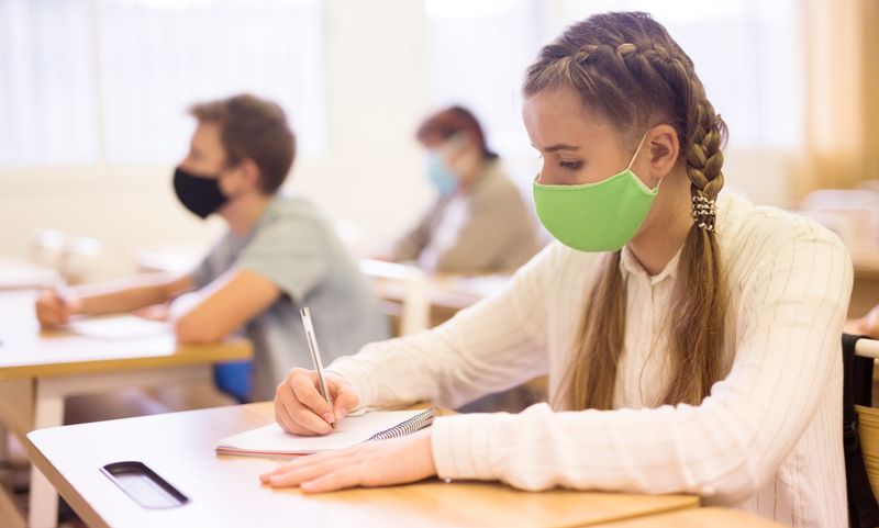 young people sat in a school classroom wearing face masks; in foreground is a girl with blonde pigtails wearing a white shirt and a green mask, writing on a notepad on the desk
