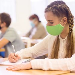 young people sat in a school classroom wearing face masks; in foreground is a girl with blonde pigtails wearing a white shirt and a green mask, writing on a notepad on the desk