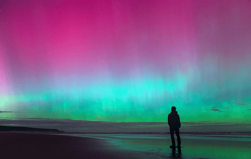 Many colors aurorae are visible in the sky over the ocean. A person is siluheatted against them
