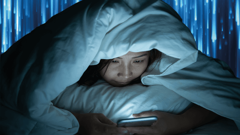 person under bed covers lying on their stomach and looking at their phone; there are blue streaks of light in the background to represent the blue light emitted from the device