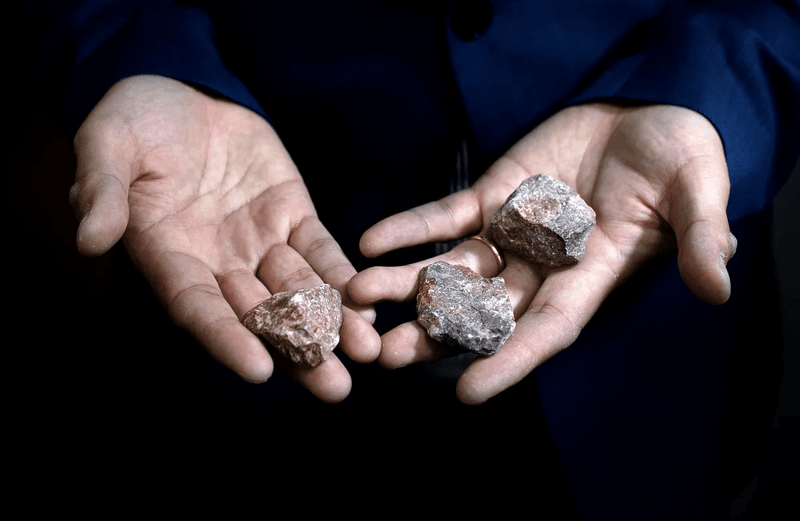 Wenhao's open palms are in sharp focus and take up nearly all of the image. The rest of Wenhao's body is barely visible. He is holding three rocks, one in his right hand, two more in his left. The rocks are a motley of reddish-pink, reddish-brown, and black colors, with some white showing wear the edges have worn.