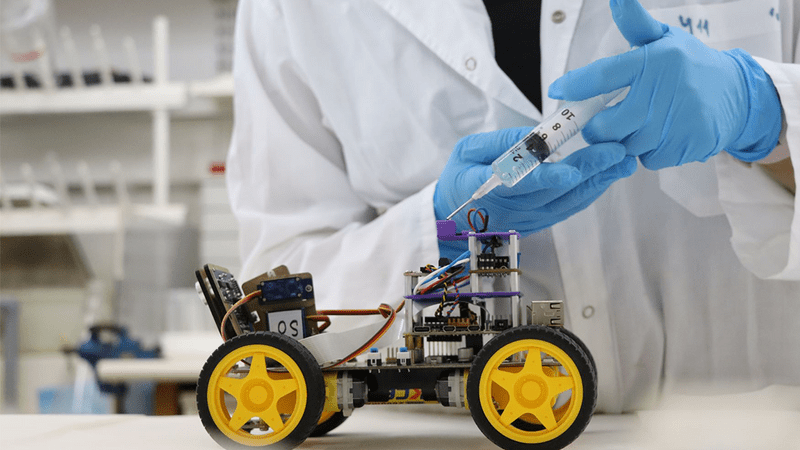 Syringe above a robot with bright yellow wheels and visible circuits and wires. Blue gloved hands are holding the syringe.