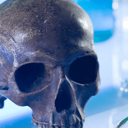 A photo of a human skull, minus the jawbone, resting on a glass table. The scene is lit to look like an advanced science lab and shows a scalpel and a brush resting next to the skull, as well as numerous bottles and beakers in the background.